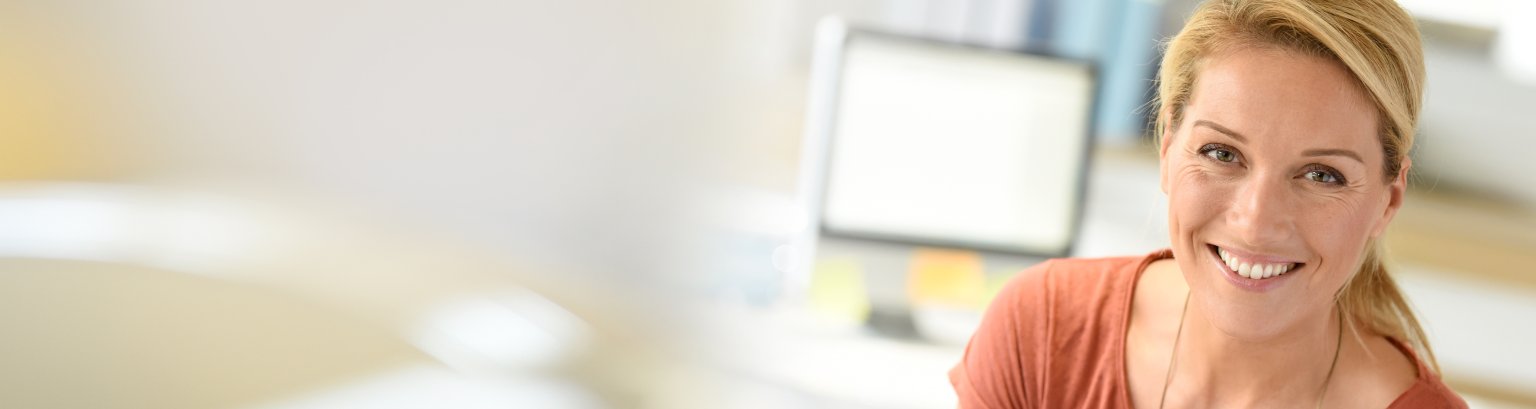 Lächelnde Frau im Büro Eine Frau am Büroarbeitsplatz, bekleidet mit einem organgen Shirt, schaut lächelnd und ausgesprochen freundlich in die Kamera.