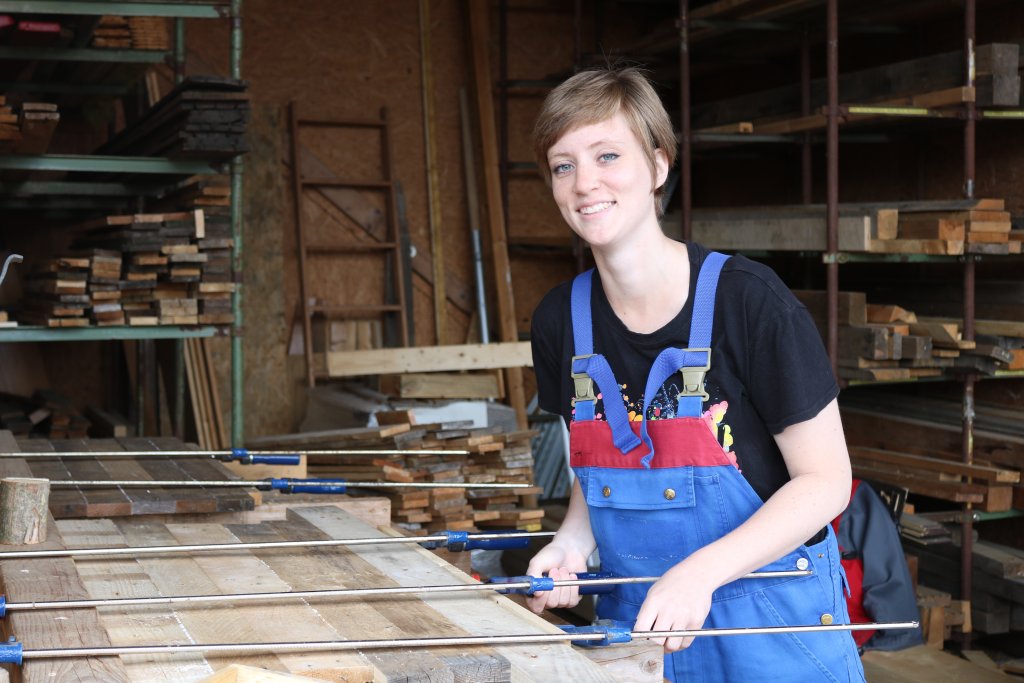 Vanessa Göcke am Arbeitsplatz Vanessa Göcke, eine junge Frau, steht in Arbeitskleidung an ihrem Arbeitsplatz in der Tischlerei und verleimt Holzleisten.