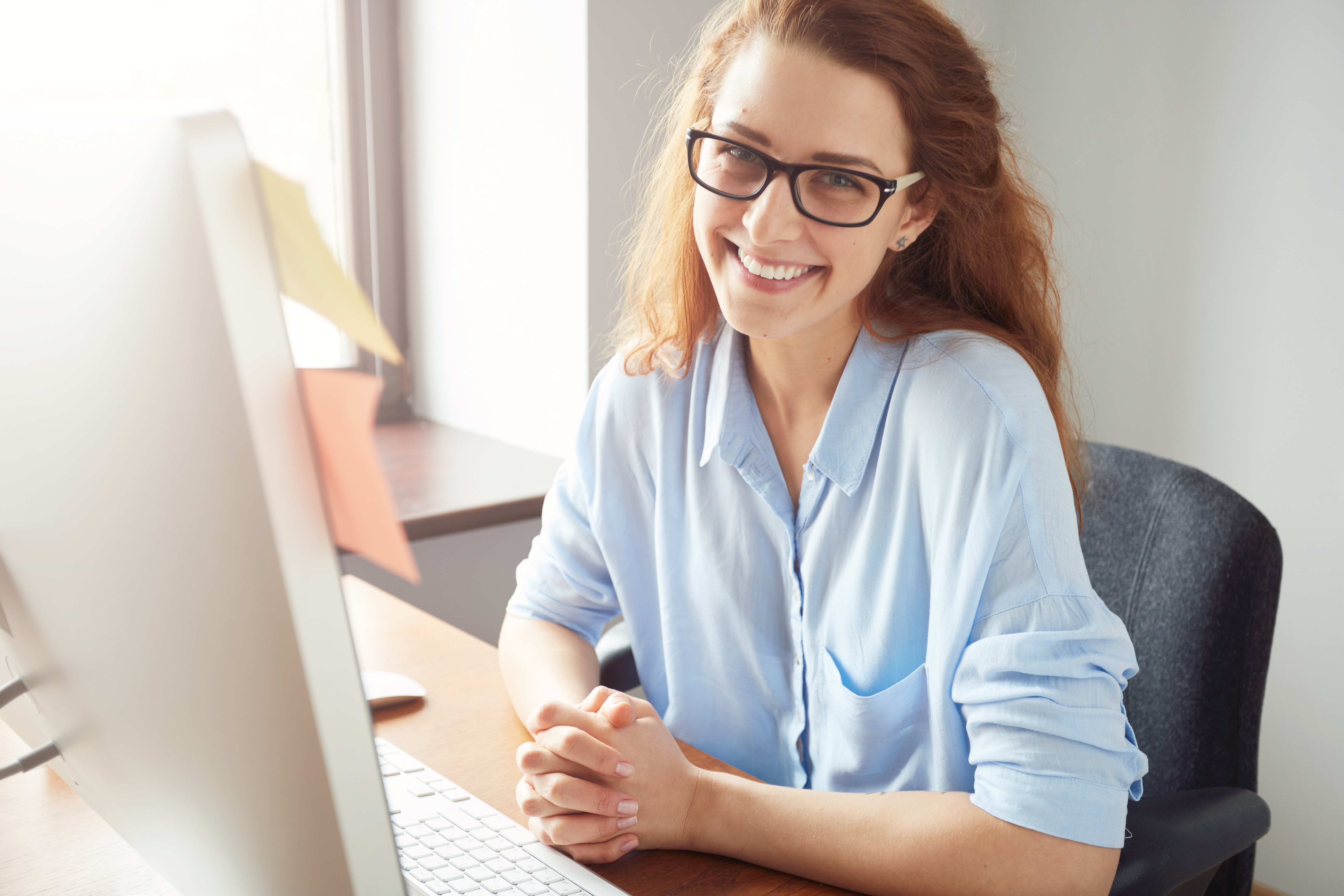 Portrait of happy successful Caucasian businesswoman in blue shirt and glasses looking and smiling at the camera. Attractive female manager sitting at desk after hard working day 