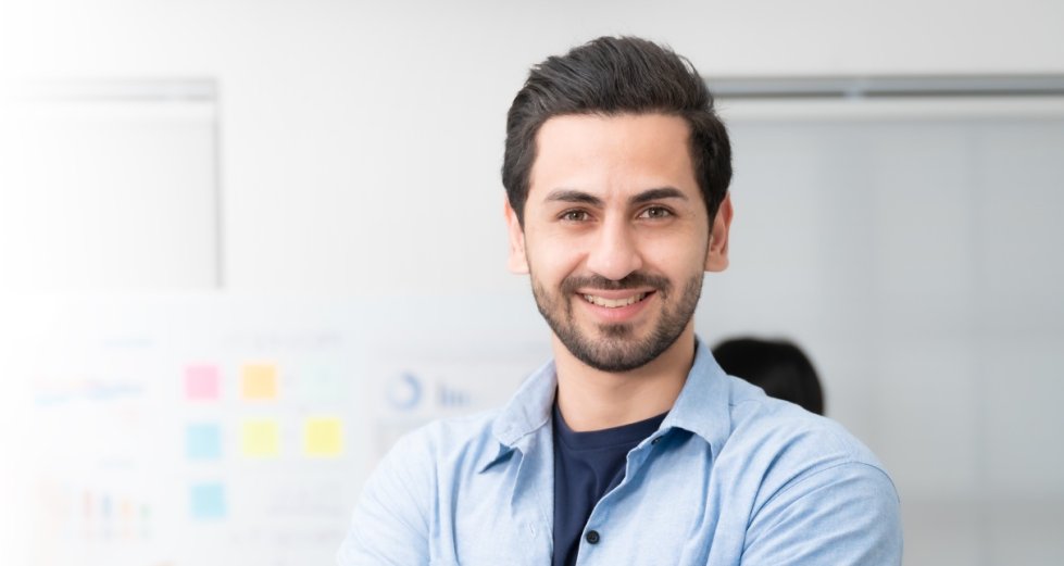 Confidence team leader. Young man owner startup business keeping arms crossed and looking at camera with smile while his colleagues working in the background Confidence team leader. Young man owner startup business keeping arms crossed and looking at camera with smile while his colleagues working in the background